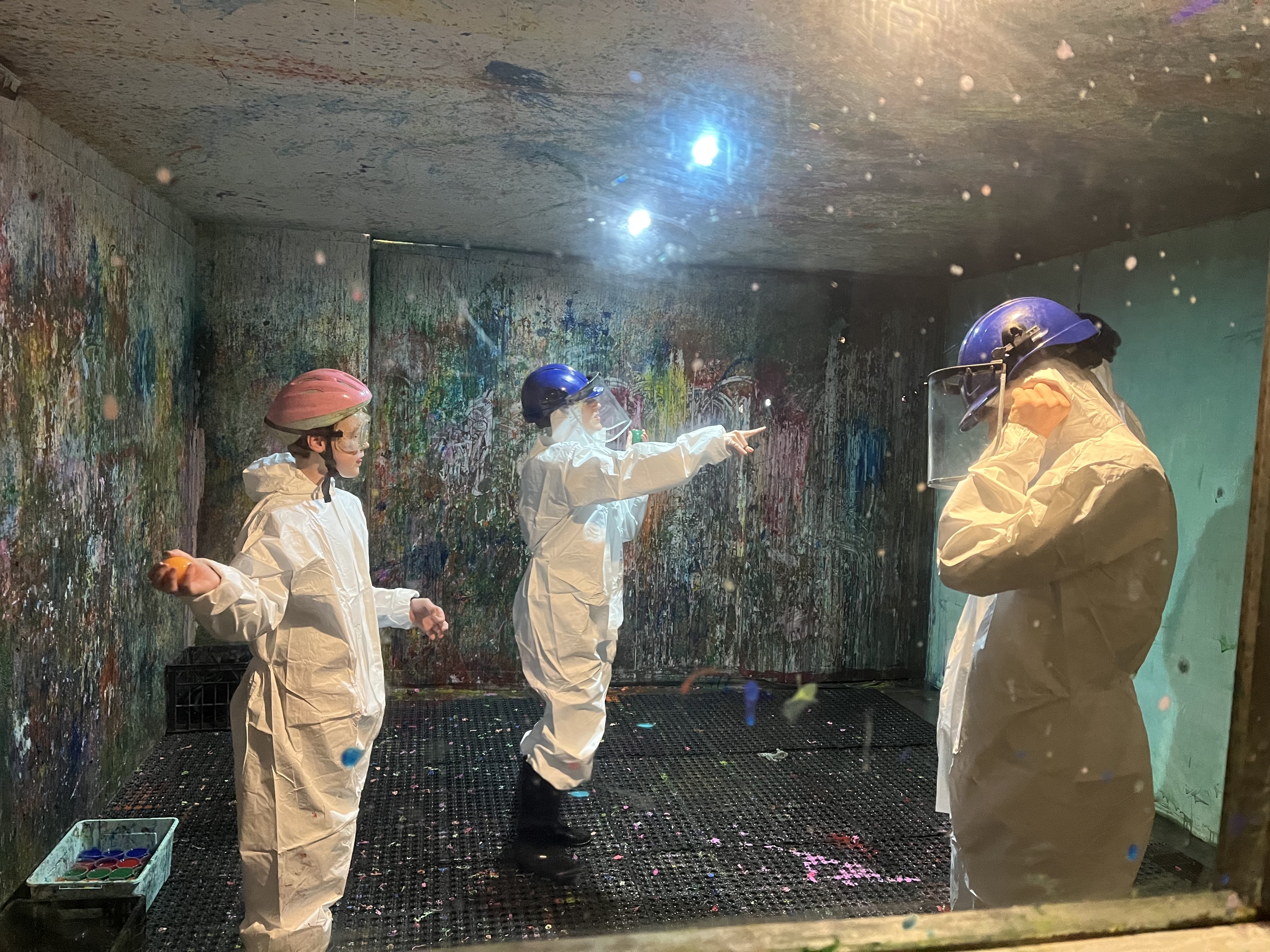 A group people wearing protective helmets and white jumpsuits are in a 'splash room' covered in paint.
