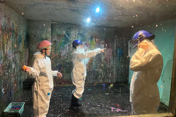 A group people wearing protective helmets and white jumpsuits are in a 'splash room' covered in paint.