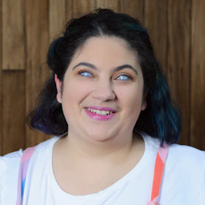 Freelance artist Olivia Muscat has short, dark brown hair. She wears vibrant makeup and rainbow-coloured overalls with white t-shirt.