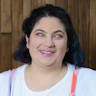 Freelance artist Olivia Muscat has short, dark brown hair. She wears vibrant makeup and rainbow-coloured overalls with white t-shirt.
