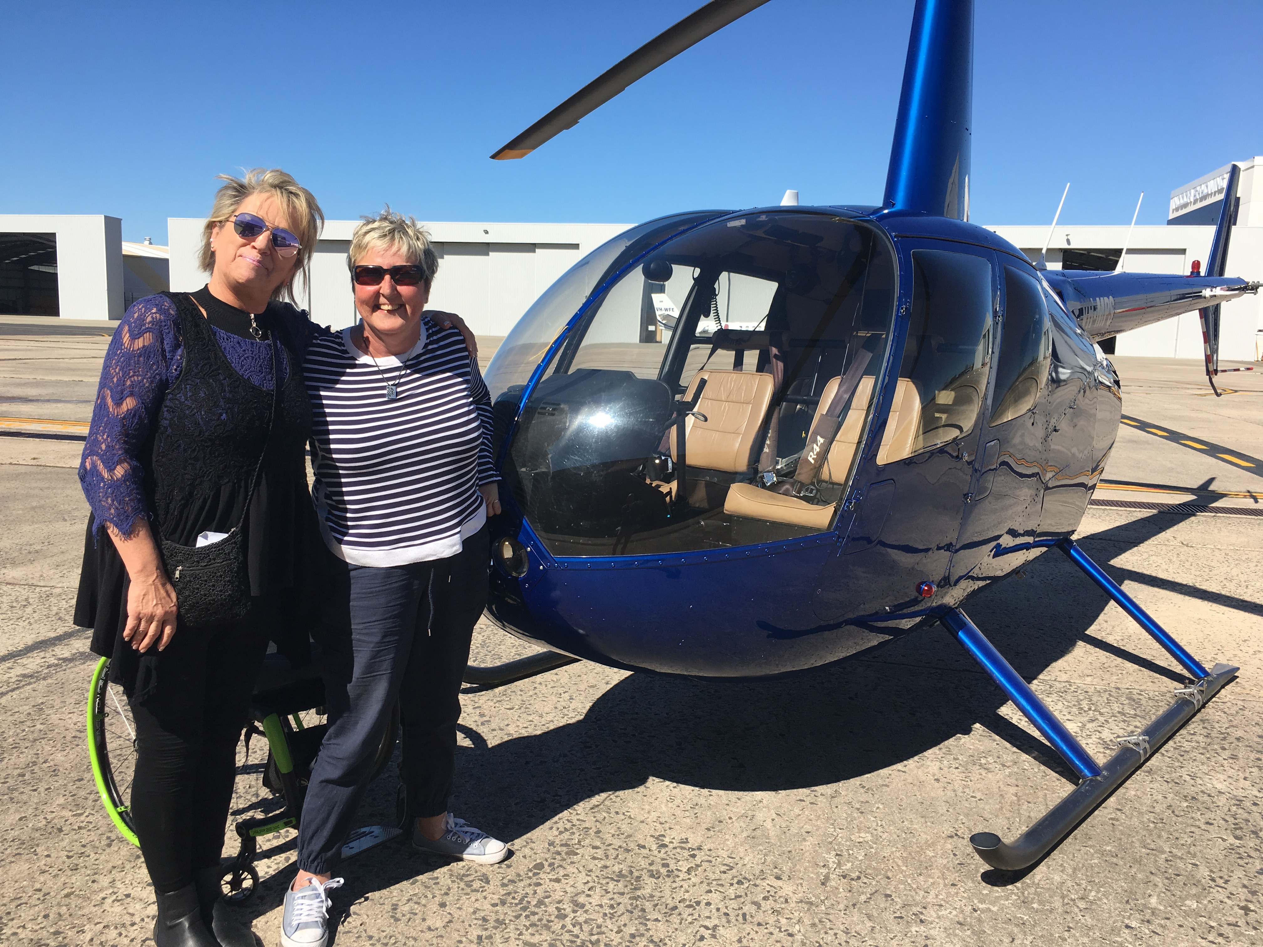 Mae, a woman with short blonde hair is wearing a navy and black outfit. Her left arm is wrapped around Christine who is wearing a stripy top. They're standing next to a helicopter in an aerodrome. A bright wheelchair is behind Christine. 