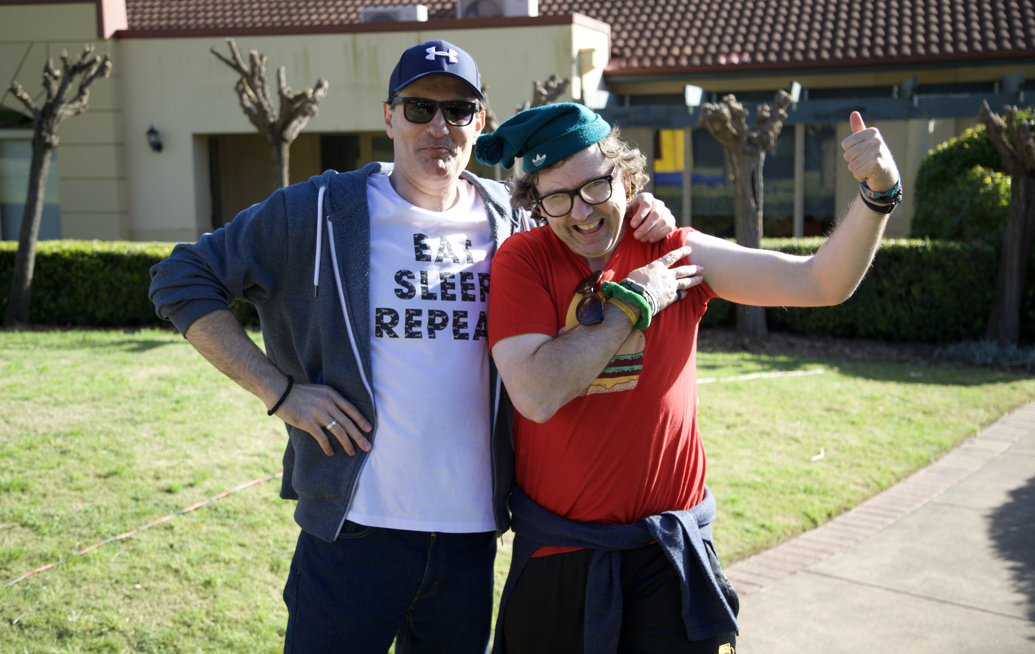 Two men are standing together smiling. One has their arm around the other. The man on the left is wearing a blue cap, blue hoodie and a white t-shirt that says 'eat sleep repeat'. The man on the right is wearing a red t-shirt, green beanie and glasses. The man on the right is flexing his left arm. 