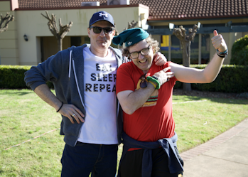 Two men are standing together smiling. One has their arm around the other. The man on the left is wearing a blue cap, blue hoodie and a white t-shirt that says 'eat sleep repeat'. The man on the right is wearing a red t-shirt, green beanie and glasses. The man on the right is flexing his left arm.