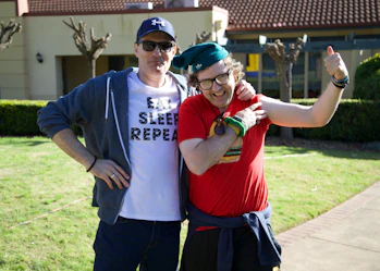Two men are standing together smiling. One has their arm around the other. The man on the left is wearing a blue cap, blue hoodie and a white t-shirt that says 'eat sleep repeat'. The man on the right is wearing a red t-shirt, green beanie and glasses. The man on the right is flexing his left arm.