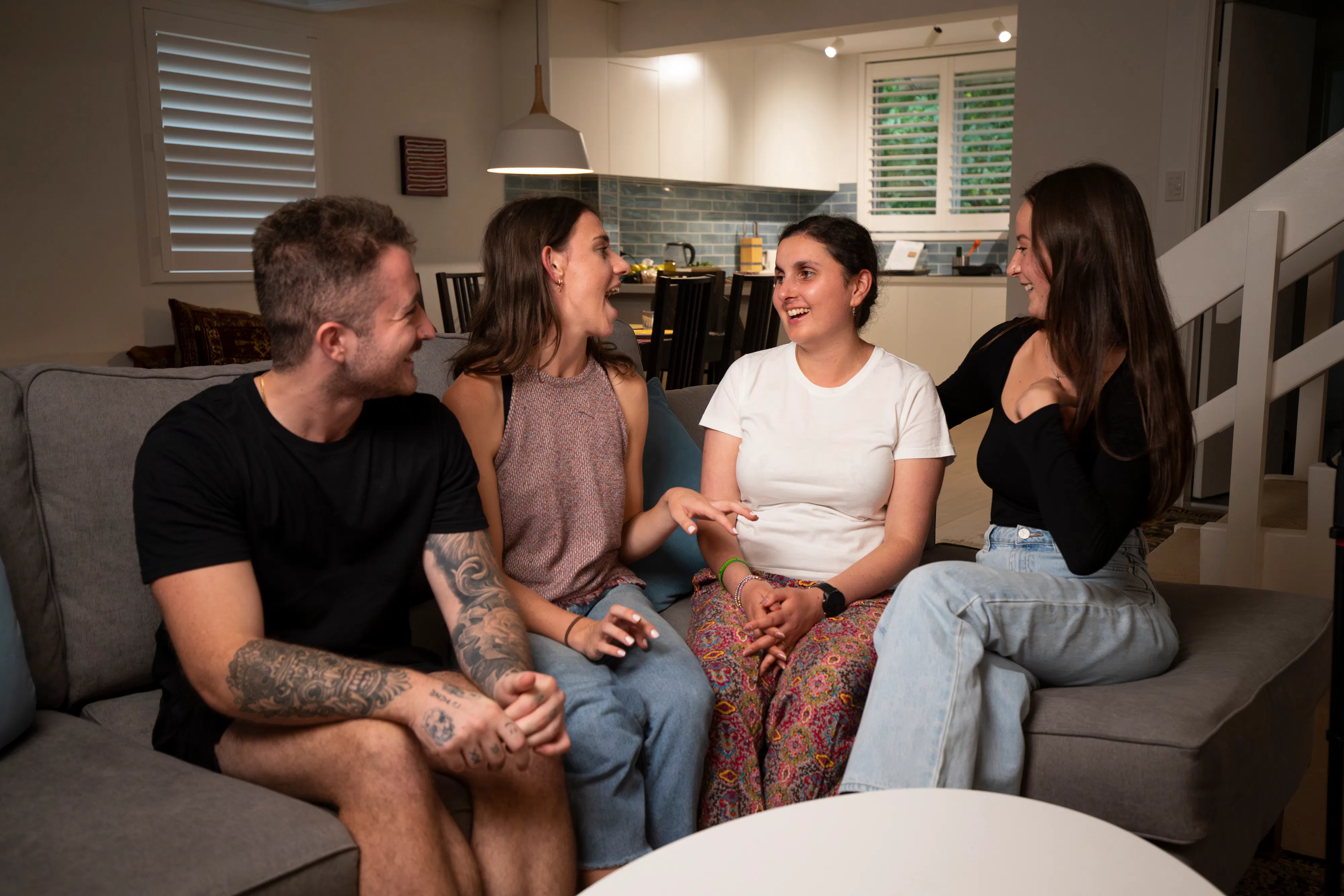 Ari and three of her support workers are sitting on a grey couch together, laughing. Ari has brown hair and is wearing a white t-shirt and long multi-coloured middle-eastern pattern pants. There is a wooden dining table and kitchen in the background.