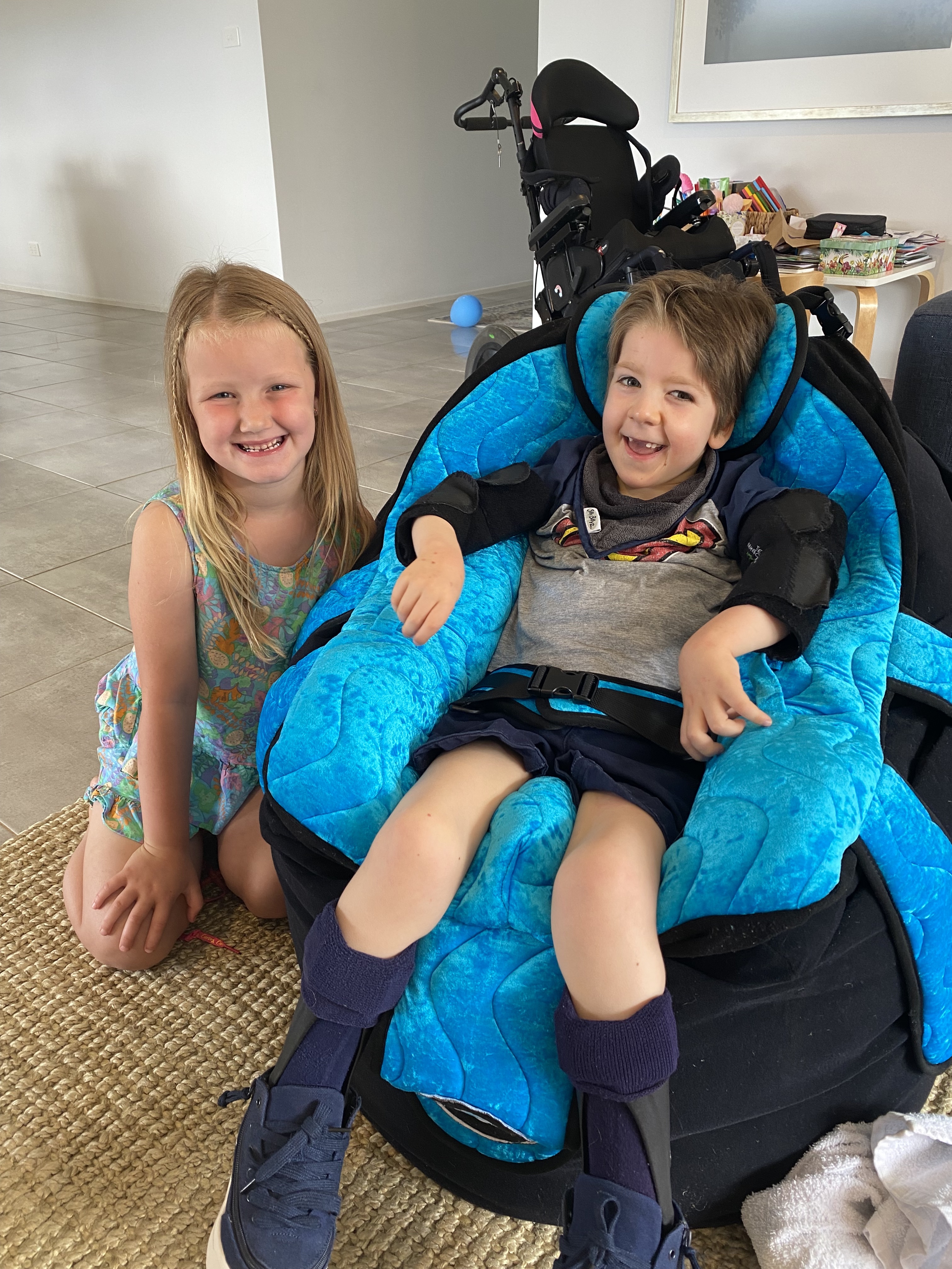 A young boy and girl - two friends - sit together in a living room smiling. The boy sits in a supportive chair. His wheelchair features in the background. 