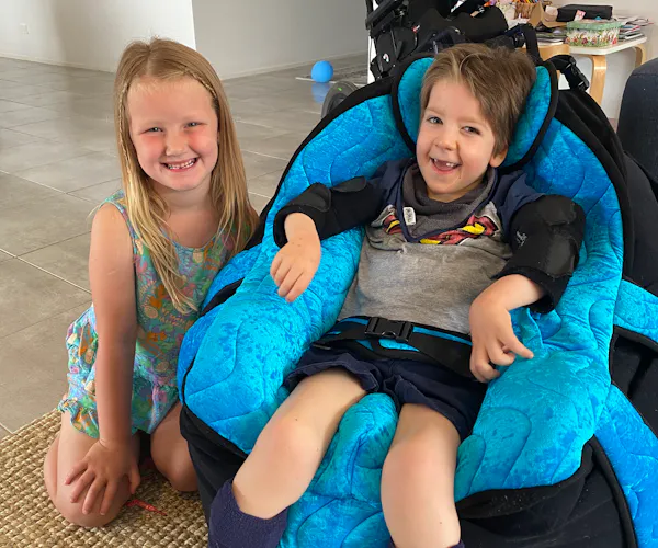 A young boy and girl - two friends - sit together in a living room smiling. The boy sits in a supportive chair. His wheelchair features in the background.
