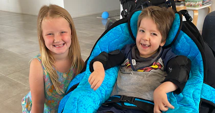 A young boy and girl - two friends - sit together in a living room smiling. The boy sits in a supportive chair. His wheelchair features in the background.