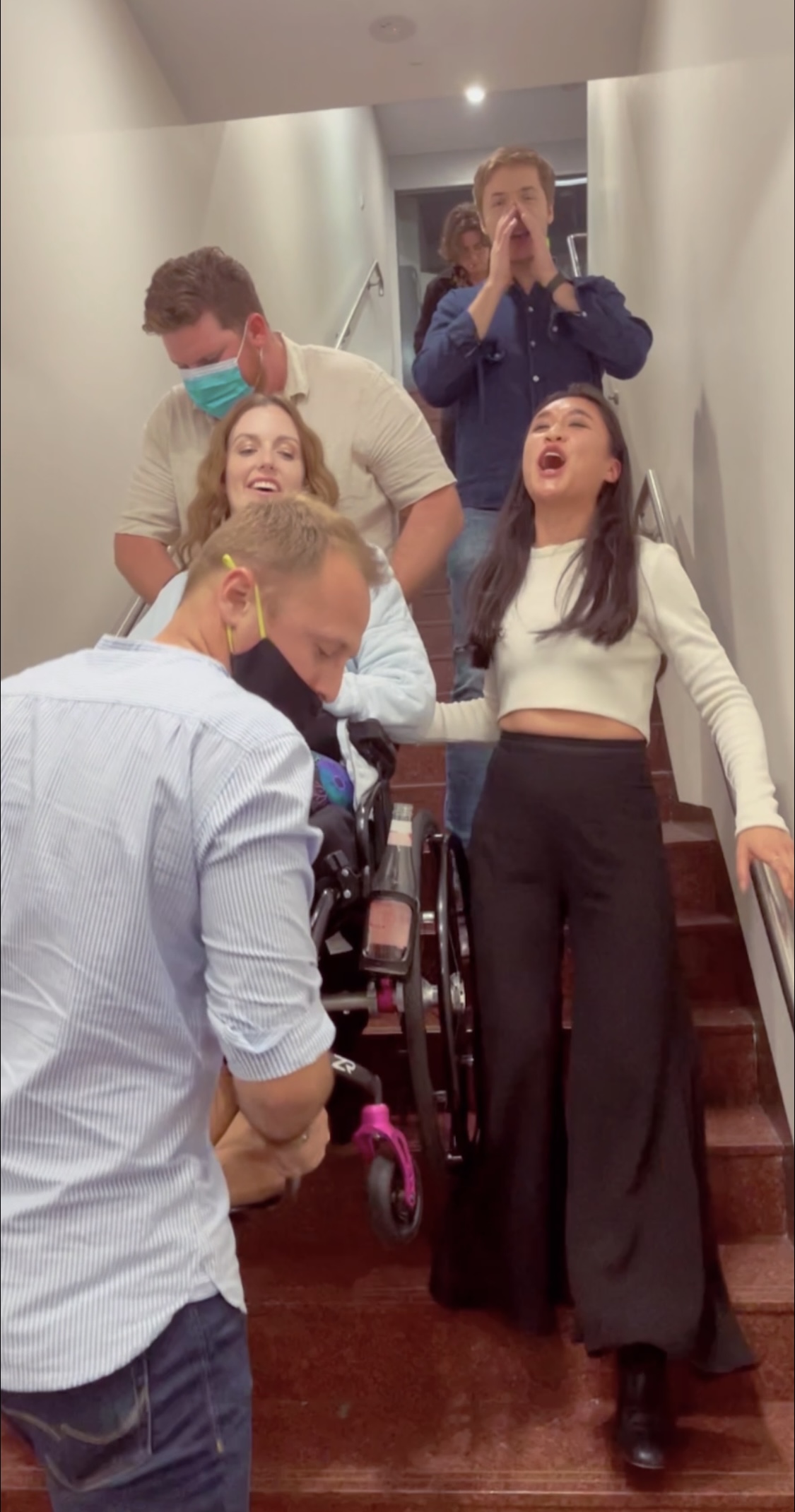 Sam has crimped hair and is smiling. She is being carried down a narrow flight of stairs by two men wearing face masks. Two of Sam's friends are going down the stairs beside Sam and are singing. Sam is a wheelchair user.  