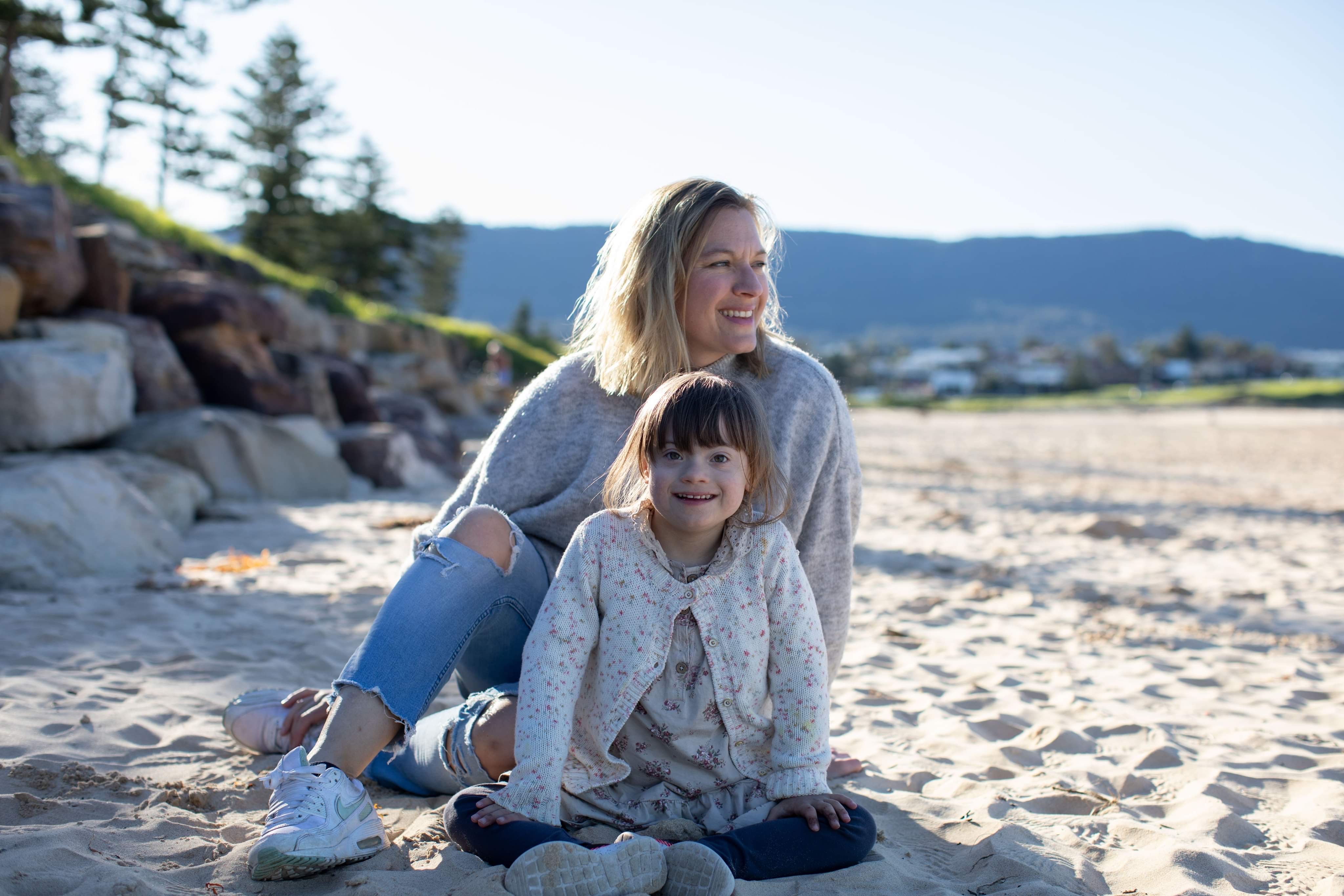 Ingerlise Svaleng, a woman with shoulder-length blonde hair, sits smiling with her young daughter, Maia, on a beach. Ingerlise wears a grey jumper, jeans and white shoes. Maia wears a floral print cardigan and dress.