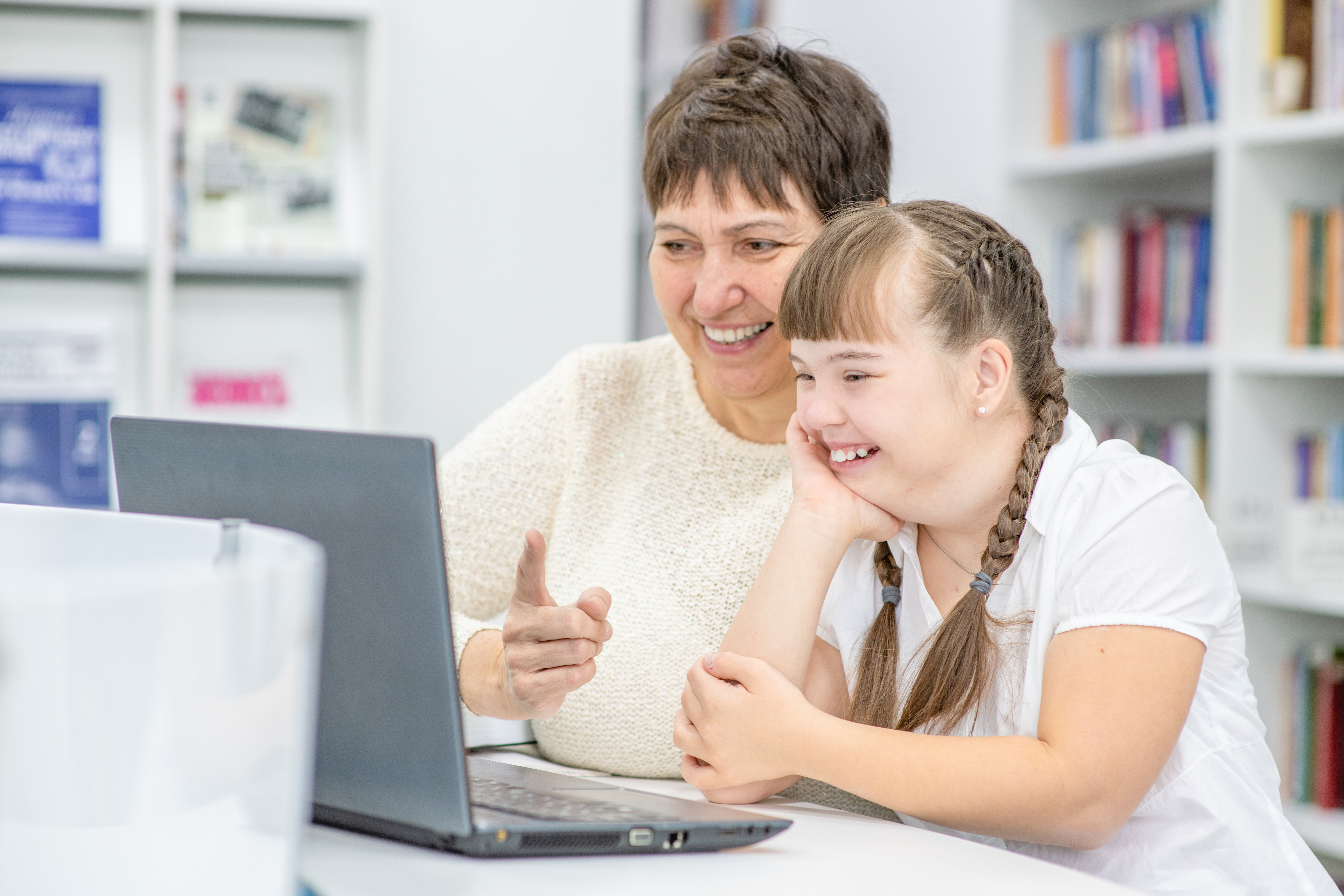 A woman and a young girl are smiling at a grey laptop. The girl has brown hair in pigtails and is wearing a white t-shirt. The woman has short brown hair and is wearing a white sweater. There are bookshelves containing books in the background.