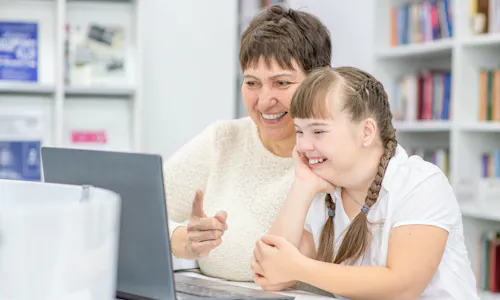 A woman and a young girl are smiling at a grey laptop. The girl has brown hair in pigtails and is wearing a white t-shirt. The woman has short brown hair and is wearing a white sweater. There are bookshelves containing books in the background.