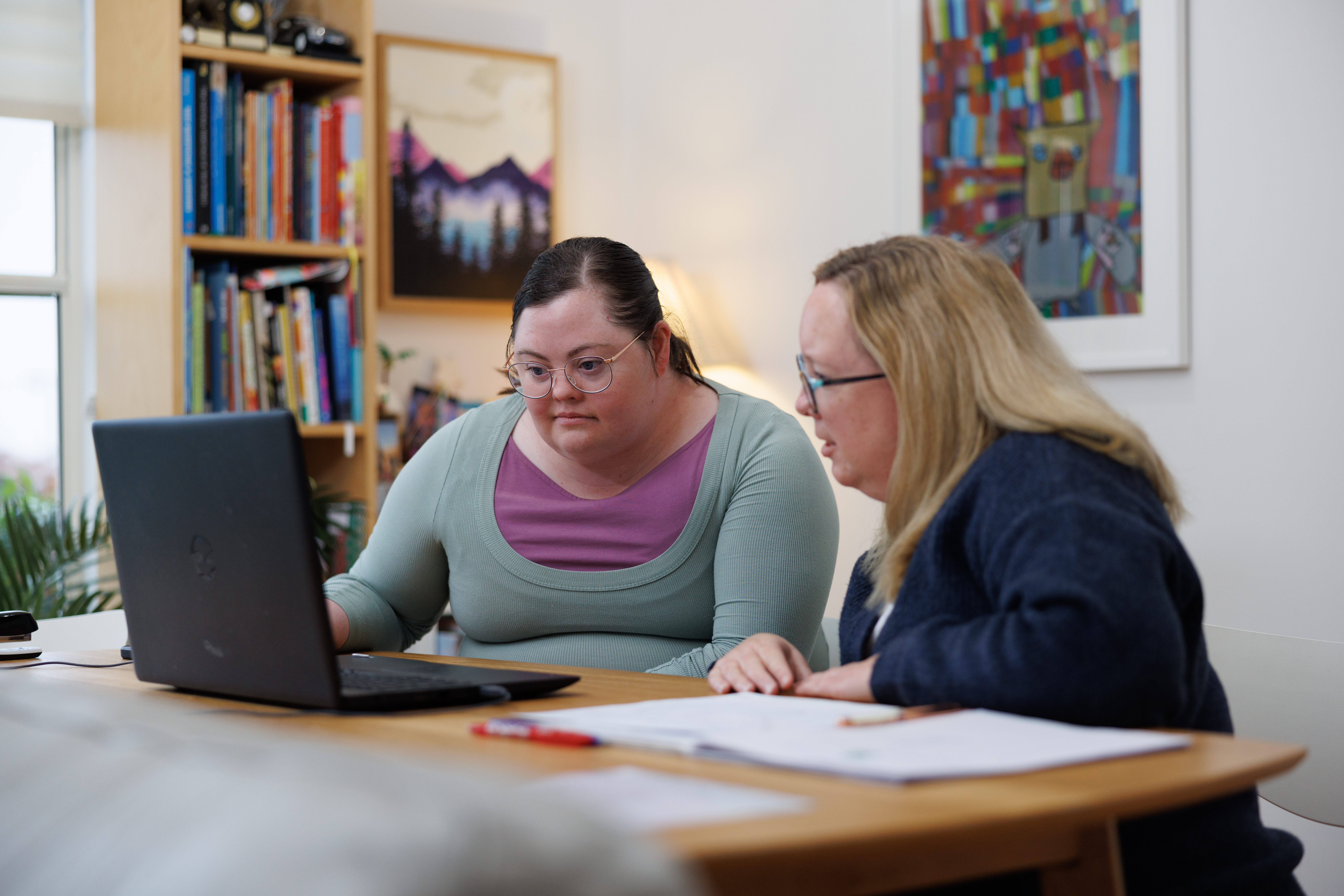 Audrey and her disability support worker, Toni, are sitting at a dining room table staring intently at a laptop. Audrey has dark brown hair pulled up into a ponytail and wears a purple and grey top. Toni has long blonde hair and wears a black jumper. They both wear glasses. Paperwork is sprawled across the table, two paintings hang on the wall and a bookshelf is filled with an array of books.