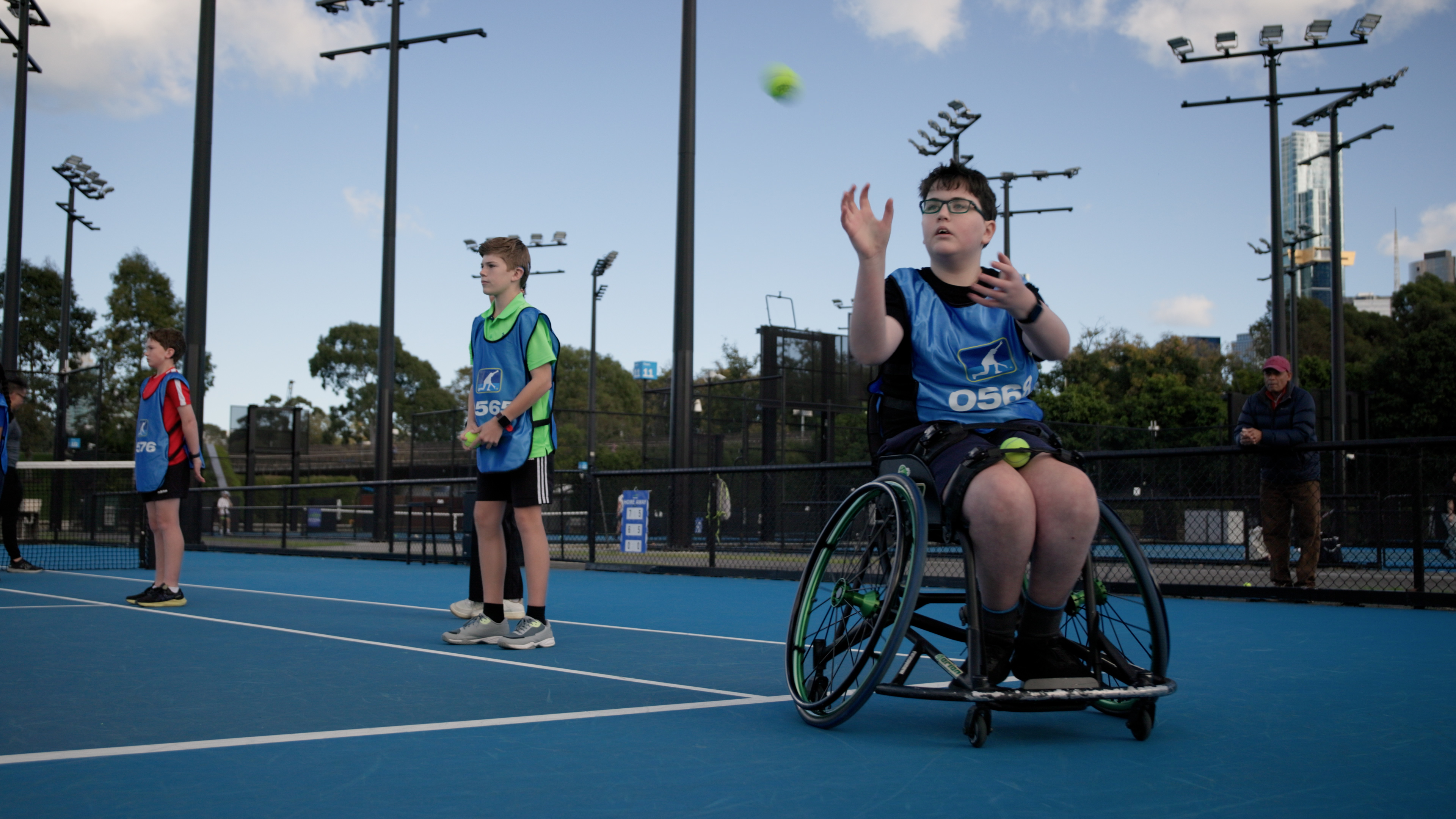 A ballkid prepares to catch a tennis ball that's whizzing through the air. He's a wheelchair user and is on a tennis court at the All Abilities Ballkids trials in Melbourne. He wears a blue smock with the word 'Ballkid' printed on it.