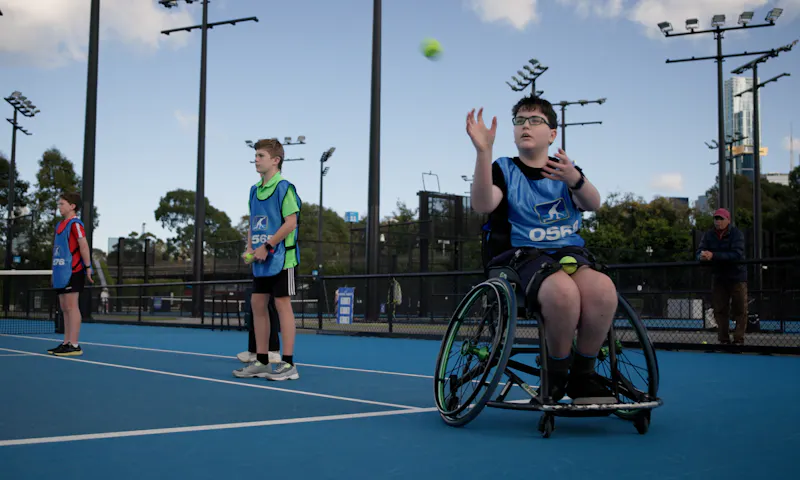 A ballkid prepares to catch a tennis ball that's whizzing through the air. He's a wheelchair user and is on a tennis court at the All Abilities Ballkids trials in Melbourne. He wears a blue smock with the word 'Ballkid' printed on it.