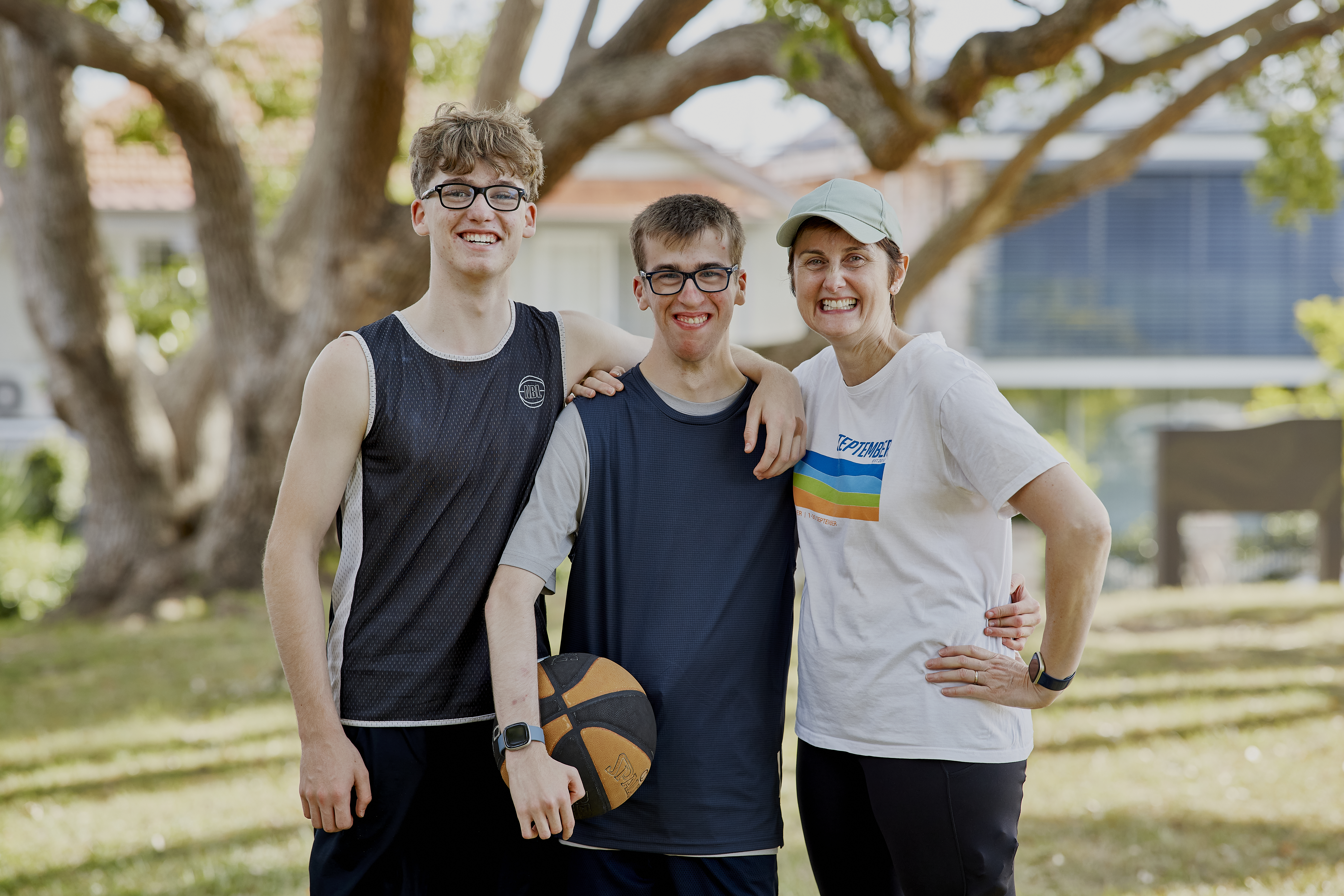 Hireup client, Brendan, is flanked by his mum and brother. They are outside and have their arms draped over his shoulders and they're all smiling. Brendan has short, brown hair and wears glasses. He's holding onto a basketball. 