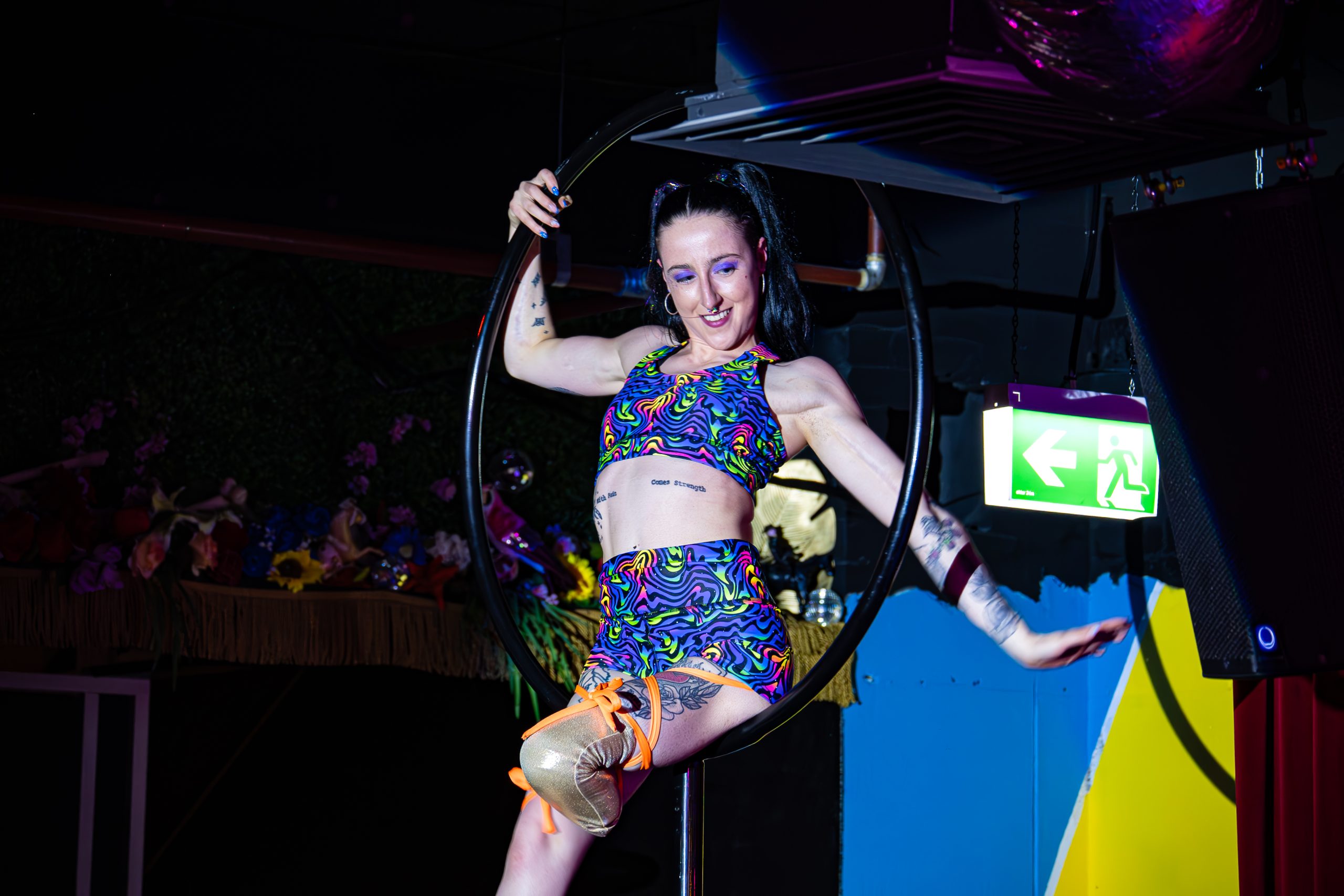 An aerialist performing in Deb Roach's show 'Criptonite' is swinging gracefully from a hoop that's attached to the ceiling. She wears a colourful cropped top and shorts, has tattoos on her arms and legs, and is a below-knee amputee. 