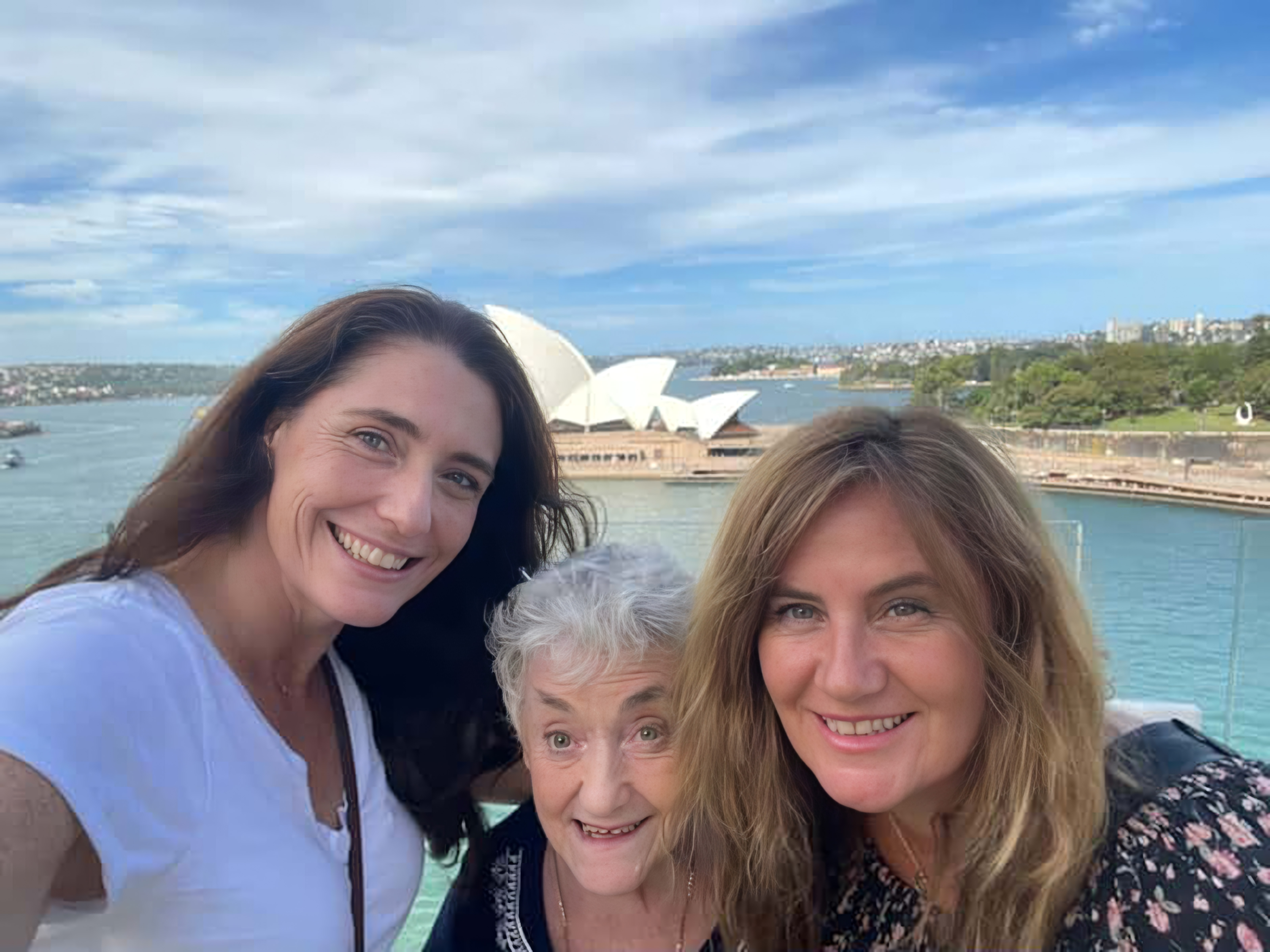Narelle and her two daughters pose in front on the Sydney Opera House on a sunny day. Narelle has white hair and is surrounded by her two daughters, they're all smiling.