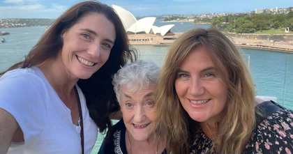 Narelle and her two daughters pose in front on the Sydney Opera House on a sunny day. Narelle has white hair and is surrounded by her two daughters, they're all smiling.