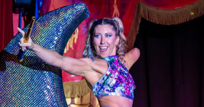 Deb Roach points into the crowd while holding onto a golden telescope. She's on the stage performing her show 'Criptonite'. Deb wears a multi-coloured sequence cropped top and sits on top of a prop that's an equally bedazzled crescent moon. Deb is an upper-extremity amputee.