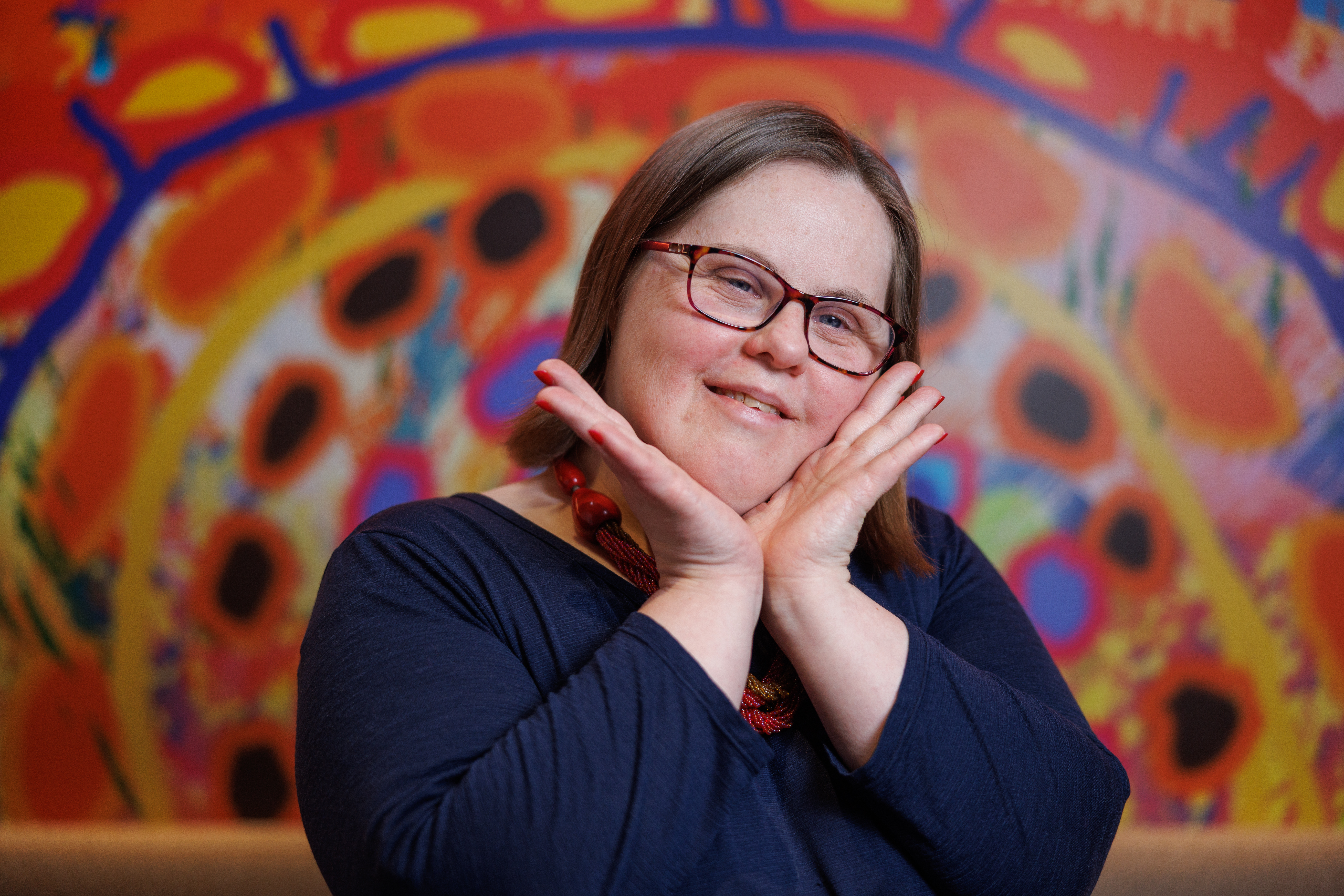 Artist Emily Crockford from Studio A poses with her hands cupping her chin, smiling. Emily has short brown hair and wears glasses. She's in front of the artwork she created for Hireup's new office. It features circles of various sizes and colours.