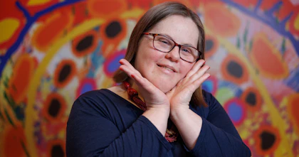 Artist Emily Crockford from Studio A poses with her hands cupping her chin, smiling. Emily has short brown hair and wears glasses. She's in front of the artwork she created for Hireup's new office. It features circles of various sizes and colours.