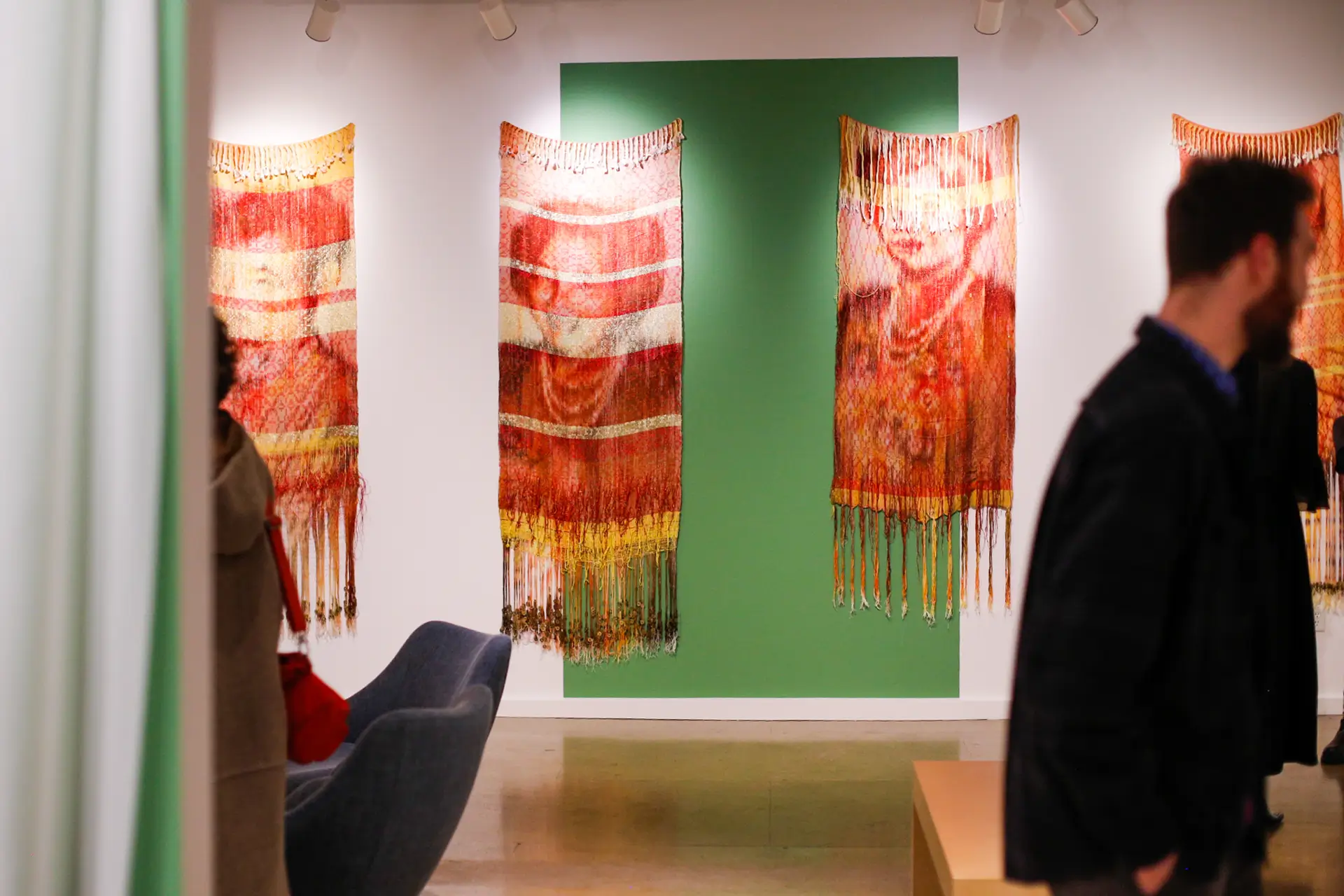 Scene from a gallery with people walking past large, woven art pieces depicting the faces of middle-aged and older women.