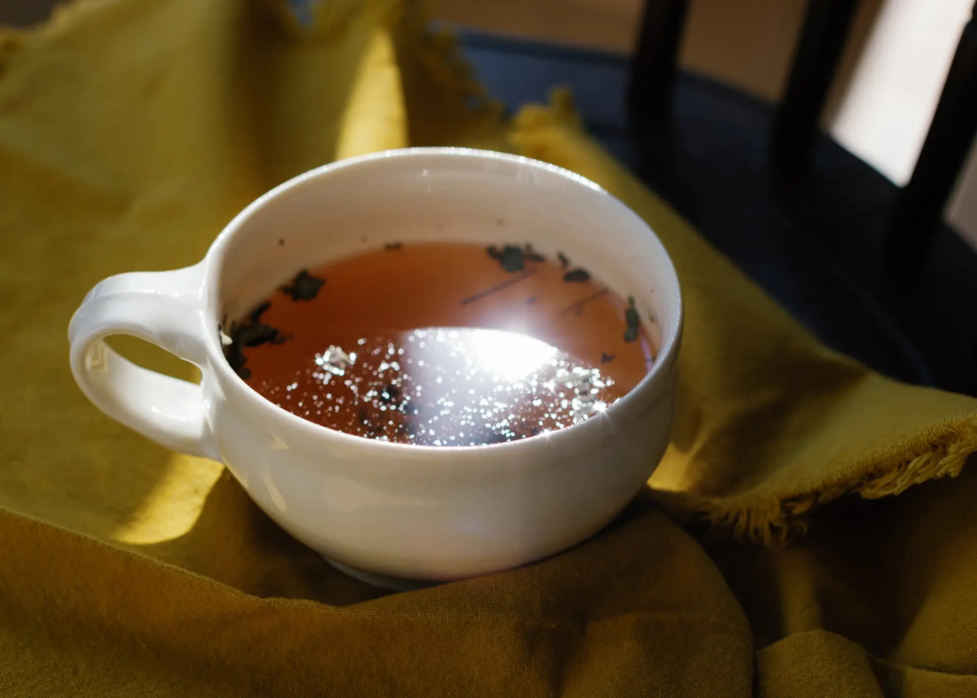 Stylized photograph of a white mug of loose leaf tea with some leaves floating in the mug and the sun reflecting off the surface of the liquid