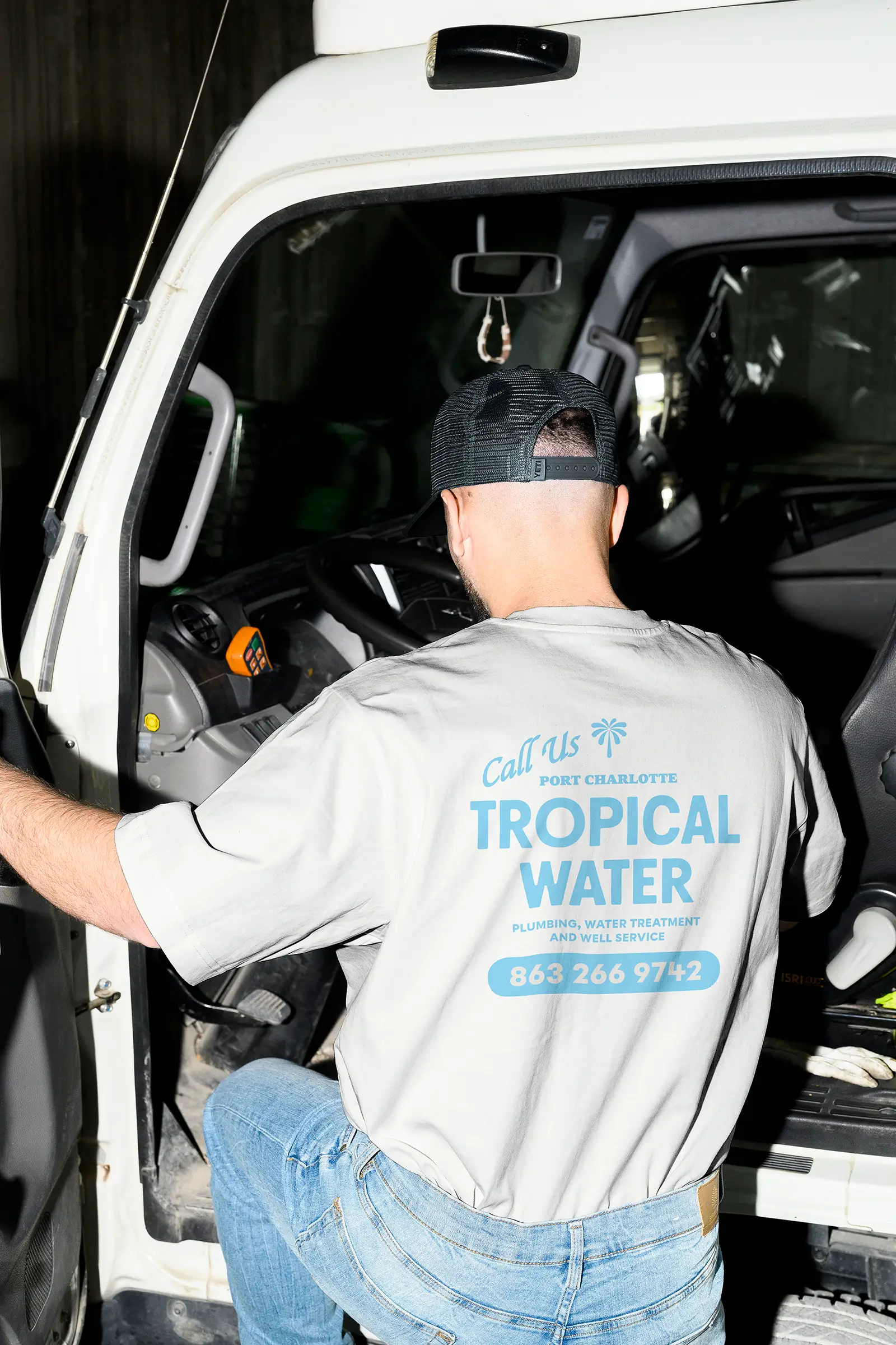 A white man in his 20s or 30s is stepping up into a white work vehicle; he's wearing a black mesh trucker hat and an off-white tee that says Tropical Water, Plumbing, Water Treatment and Well Service, 863 266 9742