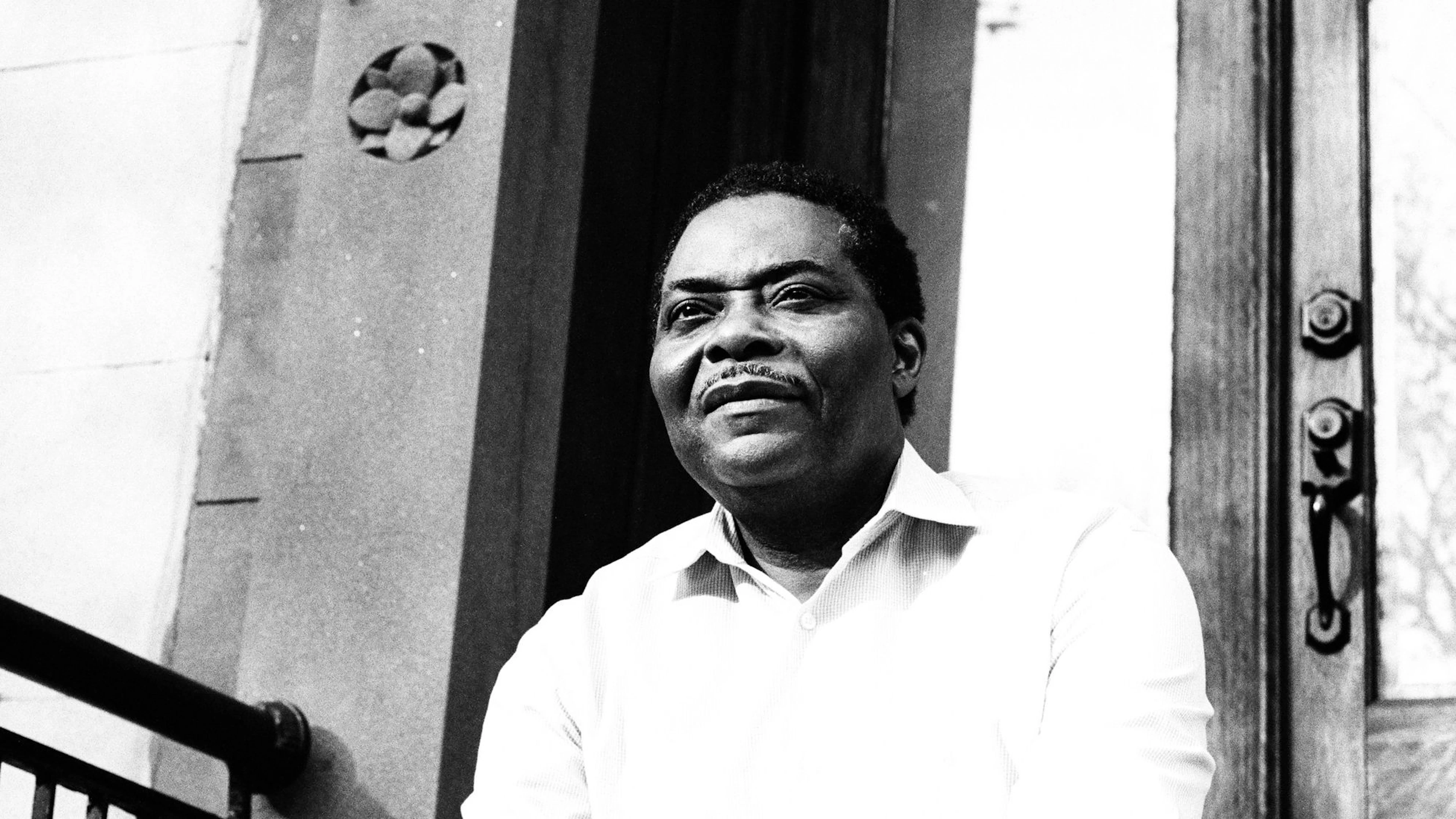 Black-and-white portrait of an older Black man seated on a brownstone stoop in a white button-down shirt, smiling softly as he looks past the camera.