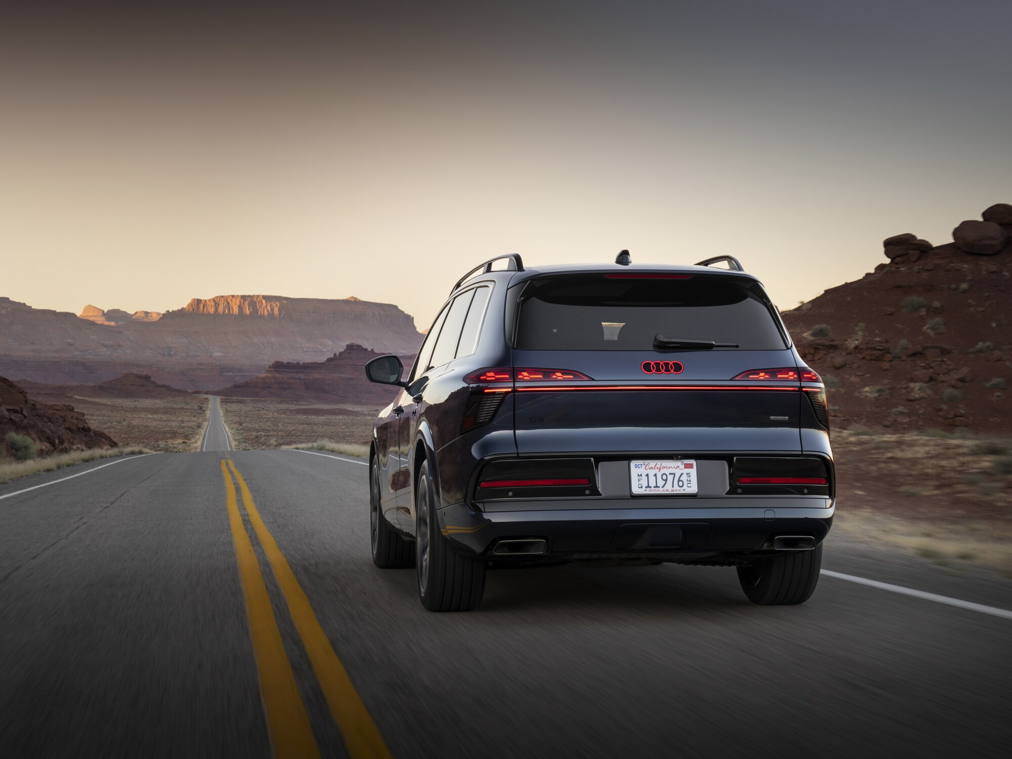 Audi Q9 in Dunkelblau, Heckansicht bei Dämmerung auf gerader Landstraße durch eine Canyon-Landschaft