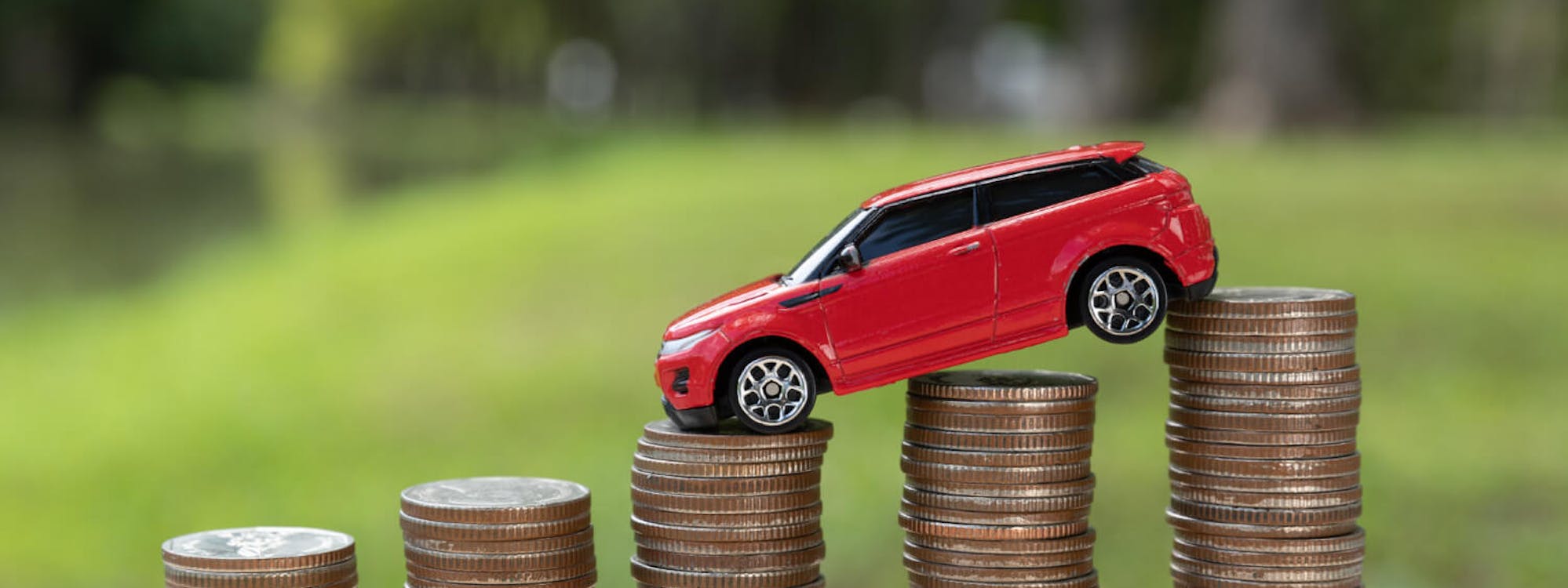 Rotes Spielzeugauto fährt eine ansteigende Reihe von Münzstapeln hinauf – symbolisch für steigenden Fahrzeugwert oder Wertverlust.