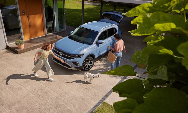 Zwei Personen und ein Hund neben einem hellblauen VW T-Cross vor einem Haus