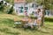 Collage avec deux images : à gauche, des meubles pliants classiques en bois au milieu d'un jardin vert et ensoleillé, une guirlande de fanions blancs au-dessus de la table et deux femmes dressant la table ; à droite, un petit banc en bois avec un coussin et une couverture gris bleu, sur lequel est posée une tasse de café et, en dessous, un chien couché sur des galets.