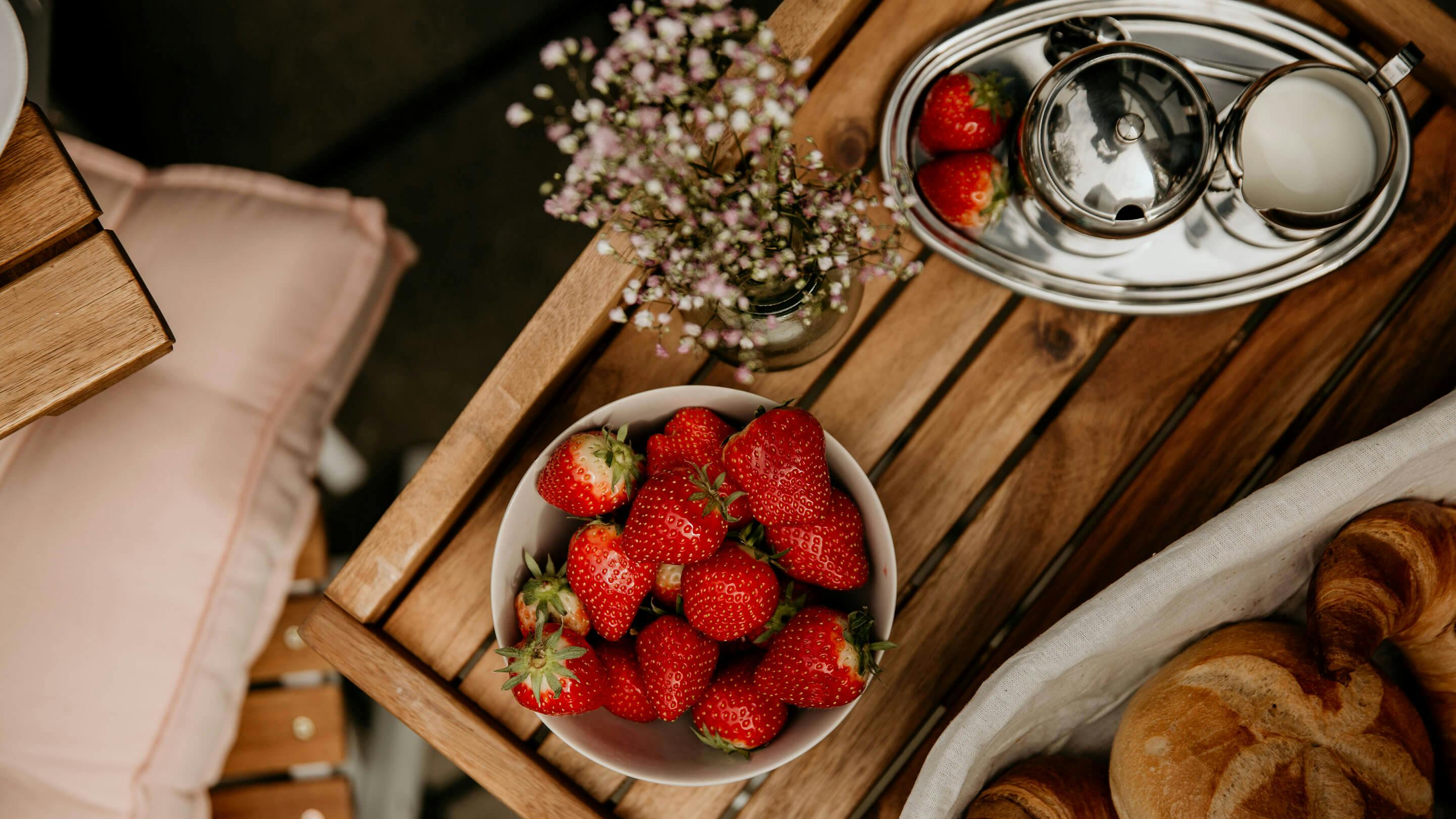 Beistelltisch aus Massivholz mit Schale mit Erdbeeren, Brotkorb mit Croissants, Vase aus Glas mit Blumen (Marke: Butlers).