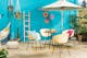 Terras met turquoise muur met een tuintafel in de industriële stijl, lichtgele kuipstoelen, een beige gebreide poef, een witte parasol, een kleurrijke hangmat en muurdecoratie, plus veel planten in bloempotten en hangende manden.