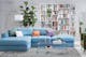 Wohnzimmer mit hellblauem Sofa, bunten Kissen, einem Glastisch sowie einem grünem Samtsessel, dazu eine Bogenlampe mit weißem Schirm, ein weißes Regal und ein schwarz-weiß karierter Teppich.