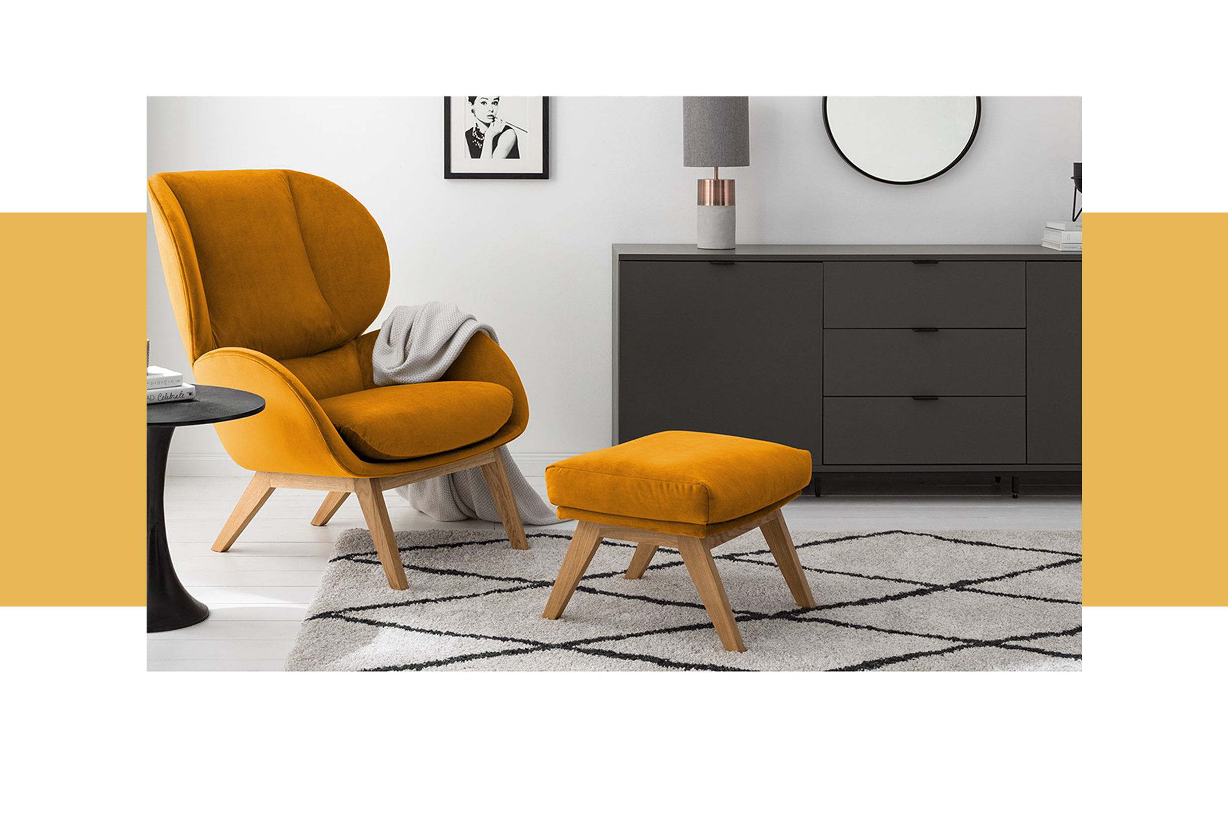 Retro-Sessel aus Samt in Orangegelb mit passendem Hocker auf einem schwarz-weißen Teppich, dazu ein schwarzer Beistelltisch, ein mattgraues Sideboard, ein Deko-Spiegel, eine Tischleuchte aus Beton sowie ein Wandbild mit Audrey Hepburn