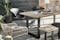 Terrasse de style industriel avec des meubles d'extérieur en acacia à la structure en métal, une chaise en rotin et d'autres chaises avec une coque noire, une table recouverte de vaisselle en céramique, de verres fumés et de planches en bois avec, en point d'orgue, une lampe d'extérieure portable.