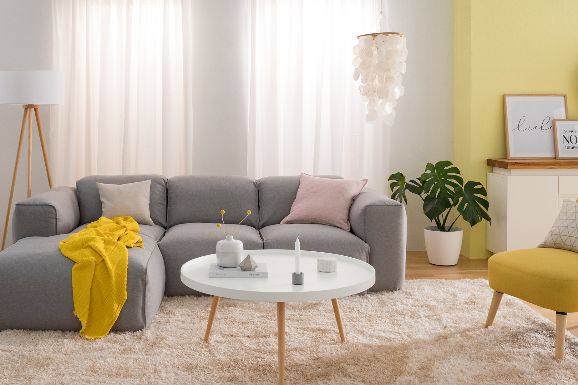 Helles Wohnzimmer mit hellgrauem Sofa, weißem Couchtisch und einer Stehlampe im Skandi-Stil, einem gelben Skandi-Sessel, einer Pendelleuchte aus Muscheln, einem weißen Sideboard und schönen Textilien.