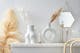 Weiße Keramikvasen in trendiger Kreis- und Po-Form sowie filigrane goldene Kerzenständer und Wandspiegel in weißer Umgebung