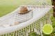 Beige hangfauteuil op een zonnig terras met planten; ernaast een foto van een grasveld met op de voorgrond een beige hangmat in boho-stijl met franjes, waarop een boek, hoed en kussen liggen.