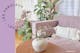 Canapé lilas avec coussins blancs de style bohème et de nombreuses plantes vertes ; à côté, un vase en verre avec des pivoines roses, derrière un papier peint à fleurs.