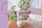 Canapé lilas avec coussins blancs de style bohème et de nombreuses plantes vertes ; à côté, un vase en verre avec des pivoines roses, derrière un papier peint à fleurs.