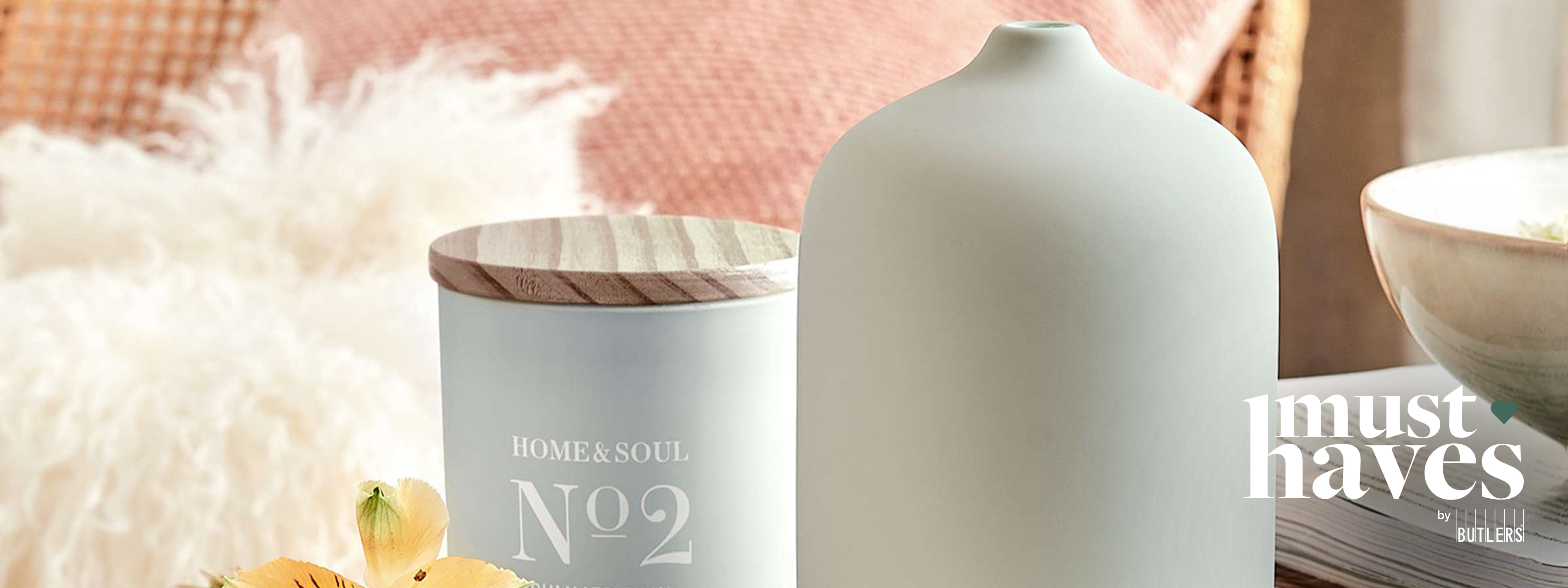 Aroma-Diffuser und Duftkerze von BUTLERS auf einer Holzbank, dekotiert mit Blüten sowie einem Schafsfell im Hintergrund.