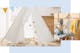 Spielecke für Kinder im Wohnzimmer, die mit einem weißen Tipi, Lichterkette, bunter Wimpelkette sowie einem weichen Kinderteppich und Kissen gestaltet ist; daneben ein grauer Polsterstuhl im Skandi-Stil, auf dem eine gelbe Kinderdecke sowie ein blaugrauer Plüschdino mit gelbem Poncho platziert ist.