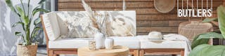 Outdoor-Loungemöbel der Serie Lexi by BUTLERS aus hellem Akazienholz mit weißen Polstern, dazu ein passender Tisch dekoriert mit weißen Vasen, ein weißer Sonnenschirm mit Fransen, ein Outdoor-Teppich und Pouf aus Jute sowie Wanddeko, Pflanzenkorb und Windlicht in Flechtoptik.