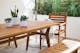 Ein Gartentisch aus Akazienholz mit weißen Pflanzentöpfen, eine passende Gartenbank sowie ein Gartenstuhl mit grauem Plaid auf einem grau-weiß gemusterten Outdoor-Teppich.