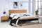 Schlafzimmer im minimalistischen Stil mit Bett, Nachttisch und Sitzbank der Serie Flox im Industrial-Stil aus Eichenholz und schwarzem Metall. Dazu schwarz-weiße Wandbilder, eine schwarze Wandleuchte, ein beiger Teppich und ein schwarzer Garderobenständer aus Metall.