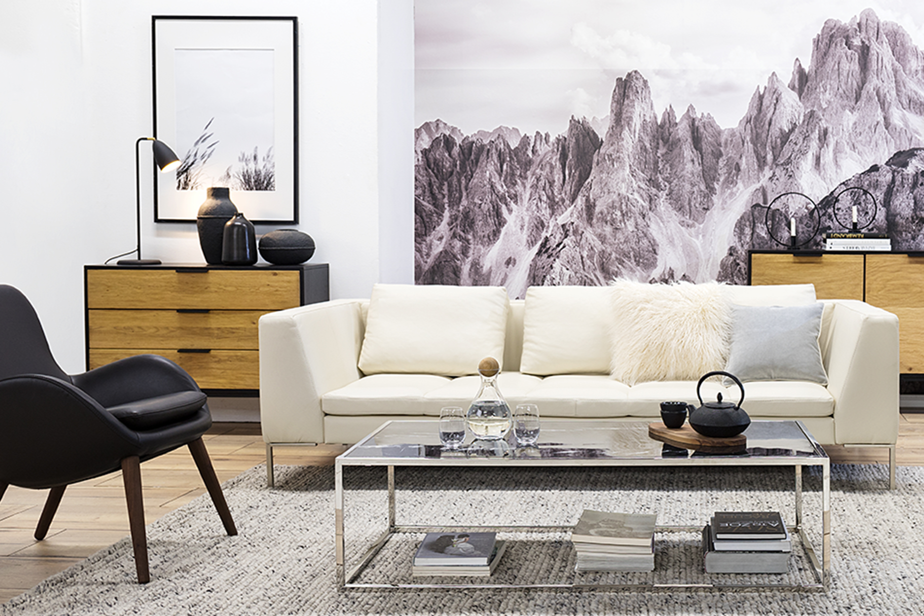 Wohnzimmer mit weißem Ledersofa und schwarzen Ledersesseln im Retro-Stil. Dazu ein silberner Couchtisch mit Glasplatte sowie eine Kommode und ein Sideboard mit Holzfronten und schwarzem Rahmen, alles vor einer Fototapete mit Berglandschaft.